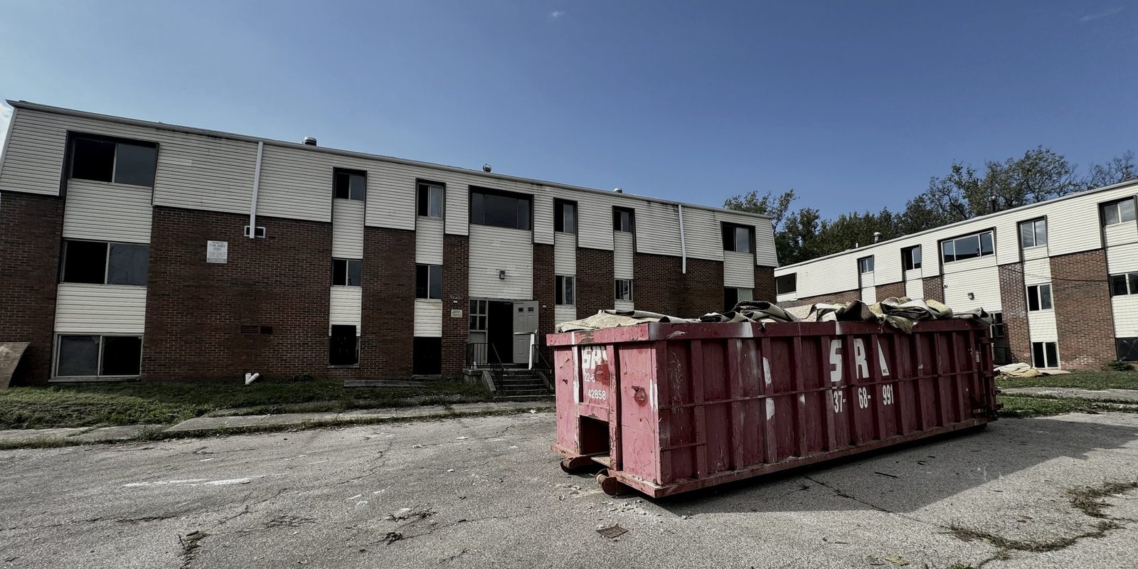 Apartment building before: boarded windows, a dumpster on a cracked empty lot